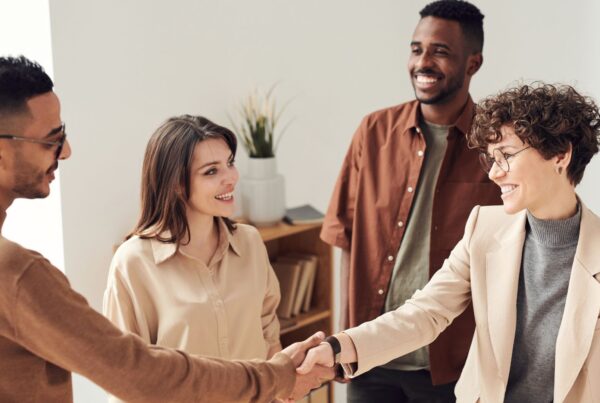 Four diverse professionals smiling and shaking hands in a bright office setting, signalling a successful business agreement.