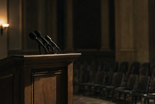 An empty wooden lectern with a cluster of microphones facing rows of vacant chairs in a dimly lit briefing room.